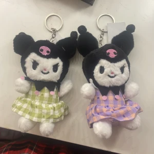 Kuromi nyckelringsväska i rutigt tyg - Supersöt Kuromi nyckelringsväska i mjuk plysch med svart huva och rosa dödskallebrodyr. Klänningen är rutigt grön eller lila med volang och små rosa knappar. Perfekt att hänga på väskan för en kawaii touch!
