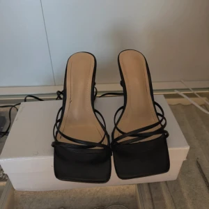 Svarta sandaler med klack och snörning -  svarta sandaler med smala remmar och fyrkantig tå. Skorna har ej så hög klack och långa snören som kan lindas runt vristen för en snygg look. Perfekta för dig som gillar minimalistisk och stilren design.