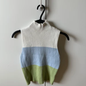 Stickad väst i blockfärger - Säljer en ribbstickad väst med hög krage i tre blockfärger: vit upptill, ljusblå i mitten och ljusgrön nedtill. Västen är ärmlös och har en clean, modern look som passar perfekt över en skjorta eller t-shirt.