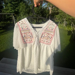 Vit broderad blus från SHEIN - Superfin vit blus från SHEIN med broderade detaljer i rosa, gult, grönt och svart på axlarna. Blusen har korta, luftiga ärmar, v-ringning med knyt och små prickar i tyget som ger en extra touch. Perfekt för dig som gillar boho-vibbar och färgglada detaljer.