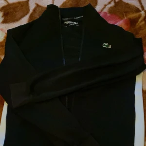 Svart Lacoste Sport hoodie XS - Svart hoodie från Lacoste Sport i klassisk passform. Tröjan har dragkedja framtill, lång ärm och den ikoniska gröna krokodilloggan på bröstet. Finns även Lacoste-tryck på bakre delen av nacken. Perfekt för en sportig och clean look.