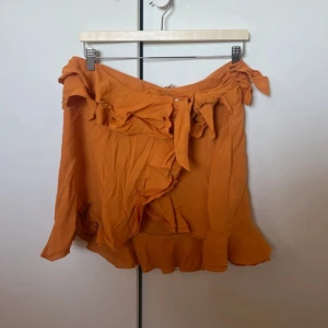 Orange omlottkjol med volang - Superfin orange kort omlottkjol med volangkant och knyt i midjan. Kjolen har en flowy känsla och är perfekt för varma dagar. Snygg att styla med en enkel topp och sneakers.