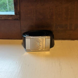 Svart skärp från Tiger of Sweden - Stilrent svart bälte från Tiger of Sweden i äkta skinn, tillverkat i Italien. Bältet har en bred, silverfärgad metallspänne med Tiger-logga ingraverad. Perfekt accessoar för att lyfta din outfit med en clean och klassisk look.