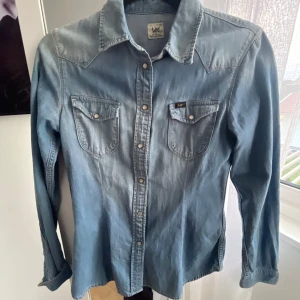Jeansskjorta Lee - Snygg blå jeansskjorta från Lee med klassisk krage och tryckknappar. Skjortan har två bröstfickor med lock och knappdetaljer. Perfekt att styla till både jeans och kjol för en cool look.