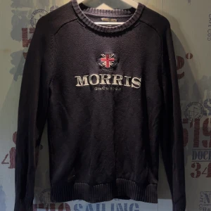 Mörkblå stickad tröja från Morris Gents - Säljer en mörkblå stickad tröja från Morris Gents med broderad logga och brittisk flagga på bröstet. Tröjan har rund halsringning och långa ärmar med ribbade muddar. Perfekt för en stilren och klassisk look.