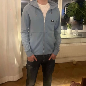Ljusblå hoodie från J.Lindeberg Storlek L - Säljer en ljusblå hoodie från J.Lindeberg med dragkedja och huva. Tröjan har långa ärmar, två fickor framtill och en broderad logotyp på bröstet. Perfekt för en avslappnad stil.