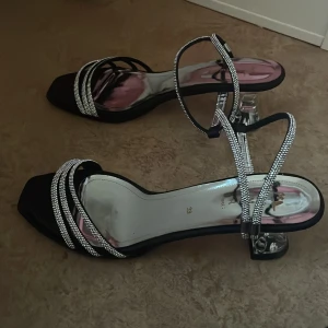 Svarta klackskor med glittriga remmar - Storlek 39, jag har storlek 38 och de passa ändå perfekt!Säljer ett par  svarta klackskor  med glittriga remmar.  Skorna har en transparent klack och tunna remmar som korsar foten. Perfekta för att lyfta din outfit med lite extra bling.