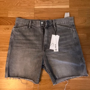 Grå jeansshorts från Zara - Säljer ett par grå jeansshorts från Zara med råa kanter och klassisk femficksdesign. Shortsen har bälteshällor, knapp och dragkedja framtill samt extra ficka på sidan. 