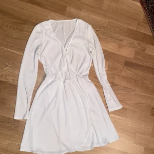 Vit klänning med lång ärm - Säljer en vit omlottklänning med långa ärmar och v-ringning. Klänningen har en lätt ribbad struktur och markerad midja som ger en feminin siluett. Perfekt för dig som gillar enkel och stilren design. Superfin till student eller ursprung!!!! 🥳🥂