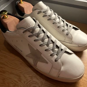 Vita sneakers från Golden Goose med grå stjärna - Snygga vita sneakers från Golden Goose med grå mockadetaljer och en ikonisk stjärna på sidan. Skorna har svarta hälpartier, grå snörning och platt sula. Perfekta för en trendig och avslappnad stil.