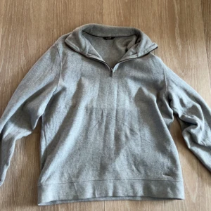 Grå halvzip tröja från Massimo Dutti - Säljer en stilren grå tröja från Massimo Dutti med halv dragkedja och hög krage. Tröjan är i mjukt material och passar perfekt till en avslappnad men snygg look. Diskret logga vid nederkanten.