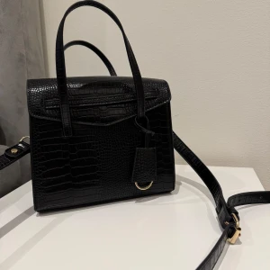 Svart axelväska i krokopräglat skinn - Snygg svart axelväska i krokopräglat skinn med både handtag och justerbar axelrem. Väskan har guldfärgade metalldetaljer och en stilren, fyrkantig form. Perfekt för att lyfta din outfit med en trendig accessoar. Helt ny utan prislapp! Aldrig använt. 🥰