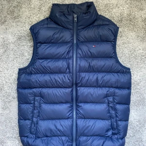 Tommy Hilfiger väst - Snygg mörkblå dunväst från Tommy Hilfiger med dragkedja framtill och två fickor. Västen har quiltad design och liten logotyp på bröstet. Perfekt för lager-på-lager och håller dig varm under kyliga dagar samt på sommaren. Västen kostar cirka 1300 kr nypris!