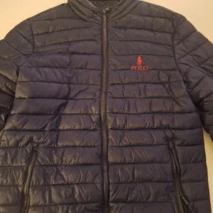 Mörkblå pufferjacka från Polo Ralph Lauren - Säljer en mörkblå pufferjacka från Polo Ralph Lauren med röd broderad logga på bröstet. Jackan har dragkedja framtill och två fickor med dragkedja. Perfekt för kyligare dagar.