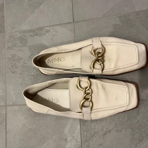 Vita loafers med gulddetaljer från MNG - Stilrena vita loafers från MNG med fyrkantig tå och dekorativa guldfärgade kedjedetaljer framtill. Skorna har låg platt sula och är tillverkade i syntetmaterial. Perfekta för en trendig och klassisk look.