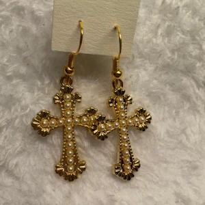 Guldiga korsörhängen med pärlor - Säljer ett par eleganta örhängen i form av kors, dekorerade med små vita pärlor. Örhängena är guldfärgade och har fina detaljer längs kanterna. Perfekta för dig som vill ha ett statement-smycke. Krokarna är i sterling silver 