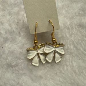 Vita rosettörhängen med gulddetaljer - Säljer ett par söta örhängen i form av vita rosetter med guldfärgade krokar och detaljer. Perfekta för att ge en gullig touch till din outfit! Krokarna är i sterling silver 