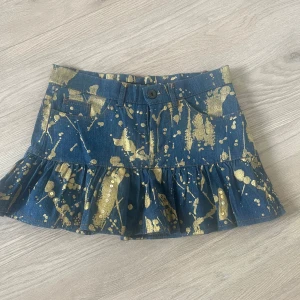 Jeanskjol med volang  - Cool jeanskjol med volangkant och unika guldfärgade stänk över hela kjolen. Kjolen har fickor både fram och bak samt knapp och dragkedja framtill. Perfekt statement-plagg för dig som vill sticka ut.