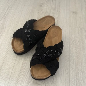 Svarta sandaler med flätade band och pärldetaljer - Säljer ett par svarta sandaler med breda, flätade band över foten och dekorativa svarta pärlor. Sandalerna har en platt sula i korkimitation och öppen tå. Perfekta för varma dagar och ger en trendig touch till outfiten. Inuikii liknande. Kommer aldrig till användning!💕
