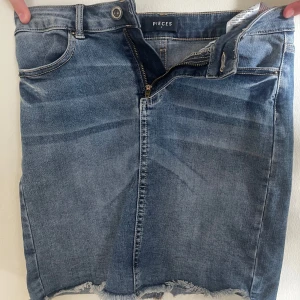 Jeanskjol från Pieces - Säljer en klassisk blå jeanskjol från Pieces med rå kant nertill och dragkedja framtill. Kjolen har normal passform och är i kort modell, perfekt till sommaren. Snygg och enkel att matcha med olika toppar.