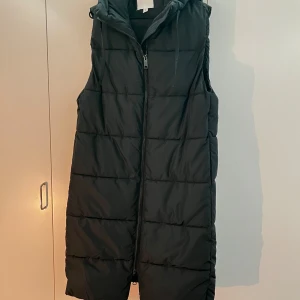 Svart lång dunväst med huva - Säljer en svart lång dunväst från H&M med huva och dragkedja framtill. Västen är quiltad och har en rak passform som går ner över knäna. Perfekt att slänga över en hoodie eller tröja för extra värme. Har inga synliga tecken på användning. Storleken är S. Priset kan diskuteras. 