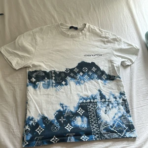 Vit och blå Louis Vuitton t-shirt - Säljer en snygg vit t-shirt från Louis Vuitton med blått batikmönster och klassiska LV-loggor. T-shirten har rund hals och korta ärmar. Märkesnamnet är tryckt på bröstet. Perfekt för dig som vill sticka ut med en exklusiv look.