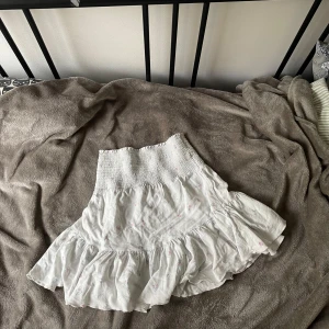 Vit blommig volangkjol med smock - Supersöt vit kjol med små blommor i rosa och blått. Kjolen har bred smock i midjan och volang nertill för en extra flowy känsla. Perfekt för varma dagar och enkel att matcha med en enkel topp. Kan även användas som en topp!