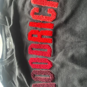 Svart t-shirt från Hoodrich med röd text - Svart t-shirt från Hoodrich med stor, broderad text i olika röda nyanser över bröstet. Klassisk passform och rund halsringning. Perfekt för dig som gillar streetwear och vill sticka ut med en snygg detalj.