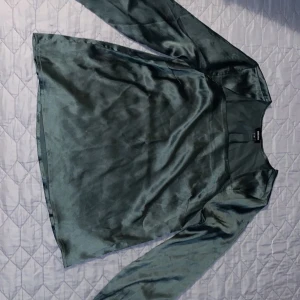 Grön satinblus med långa ärmar - Säljer en elegant grön blus i glansigt satinmaterial. Blusen har långa ärmar och en rak passform, perfekt för att lyfta vilken outfit som helst. Superfin färg och mjukt tyg!