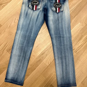 Blå jeans från Ecko Unltd med broderade bakfickor size 32-34 - Säljer ett par blå jeans från Ecko Unltd med raka ben och snygga slitningar. Bakfickorna har broderade loggor och ränder i svart, vitt och rött. Klassisk femficksmodell med knappgylf och bälteshällor.