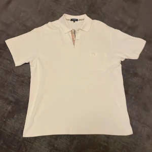 Vit piké från Burberry - Säljer en klassisk Beige pikétröja från Burberry med kort ärm och ikoniskt rutigt tyg på insidan av knapplisten. Tröjan har en bröstficka och stilren design, perfekt för en clean och snygg look.