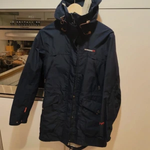Mörkblå vindjacka från Didriksons - Säljer en mörkblå vindjacka från Didriksons med hög krage och justerbar huva. Jackan har dragkedja framtill, knappar och flera fickor. Perfekt för blåsiga dagar och har en sportig look. Endast använd någon enstaka gång.