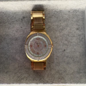 Guldig klocka med stenar från Regal - Säljer en snygg guldig klocka från Regal med glittriga stenar runt urtavlan. Klockan har en metall-länk och tydliga siffror på urtavlan. Perfekt accessoar för att lyfta din stil.