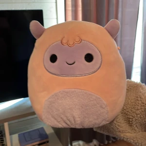 Squishmallows gosedjur lila och beige - Supermjuk och rund Squishmallows gosedjur i lila och beige med sött ansikte och små öron. Perfekt som kramkompis eller dekoration. Märkt med original Squishmallows-tag.