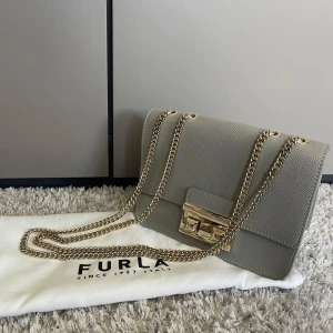 Grå axelväska från Furla med guldkedja - Najs väska från furla med guld detaljer, med nitar på framsidan. Går att bära på två sätt, man kan justera kedjan. Nypris ca. 2900kr (tillkommer dustbag och aldrig använd)
