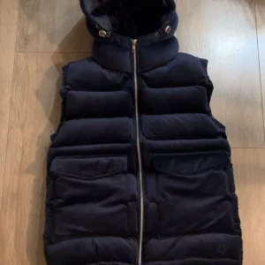 Svart dunväst med huva från Vendiera - Säljer en mörkblå dunväst från Vendiera med huva och dragkedja framtill. Västen har två stora fickor framtill och är quiltad för extra värme. Perfekt för lager-på-lager under kyliga dagar.