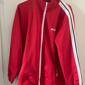 Röd track jacket från Palm Angels - Röd track jacket från Palm Angels med vit dragkedja och vit rand med svart detalj längs ärmarna. Jackan har hög krage och ribbade muddar. Palm Angels-logga på bröstet. Perfekt för en sportig och trendig look. Har byxor till denna tsm 1200kr 