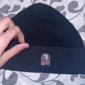 Svart ribbad mössa från Parajumpers - Säljer en svart ribbstickad mössa från Parajumpers med klassisk logotyp framtill. Mössan är enkel och stilren, perfekt för kyliga dagar. Tillverkad i mjukt material som sitter skönt på huvudet. Endast använd fåtal gånger, i skick som en ny.