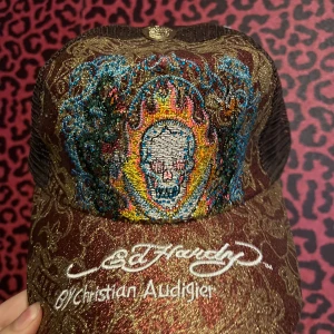 Brun Ed Hardy keps med broderad döskalle - Säljer en brun och guldglittrig keps från Ed Hardy med broderad döskalle i färgglada nyanser framtill. Kepsen har nät på sidorna och bak, samt vit brodyr med logga och Christian Audigier-text framtill.