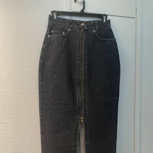 Svart jeanskjol med dragkedja - Säljer en svart midi jeanskjol med hög midja och hel dragkedja framtill. Kjolen har klassiska jeansfickor både fram och bak samt en liten slits framtill. Perfekt till sneakers eller boots.