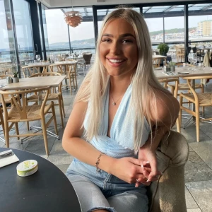 Ljusblå omlottopp med knyt - Säljer en ljusblå topp med knyt i nacken och djup v-ringning. Toppen har en lätt glansig yta och är ärmlös, perfekt för varma dagar. Snygg och trendig modell som passar till många olika outfits.