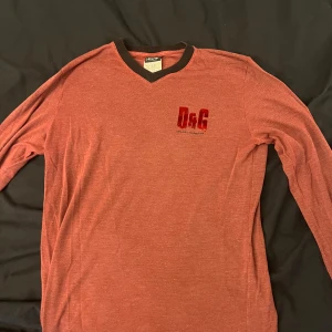 Röd långärmad tröja från Dolce & Gabbana - Säljer en röd långärmad tröja från Dolce & Gabbana med svart v-ringad krage och D&G-logga på bröstet. Perfekt för dig som gillar stilrena och klassiska plagg med en twist. Pris kan förhandlas vid snabb affär
