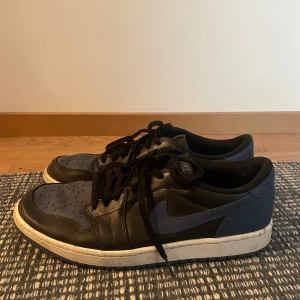 Jordan 1  low  - Säljer ett par Nike Air Jordans Low i svart och mörkblått med klassisk swoosh på sidan. Skorna har snörning, rund tå och platt vit sula. Perfekta för dig som gillar streetwear och vill ha en ikonisk sneaker i garderoben.