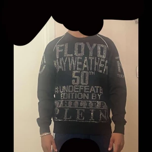 Svart sweatshirt från Philipp Plein Floyd Mayweather Edition - Cool svart sweatshirt från Philipp Plein med stora silvriga tryck och texten 'Floyd Mayweather 50th Undefeated Edition'. Tröjan har rund halsringning och långa ärmar. Perfekt för dig som gillar statement-plagg och exklusiva samarbeten.