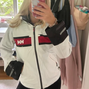 Vit vindjacka från Helly Hansen - Säljer en vit vindjacka från Helly Hansen med svarta och röda detaljer samt dragkedja framtill. Jackan har hög krage och luva . Perfekt till skärgården och båt. Supersnygg och i jättebra skick