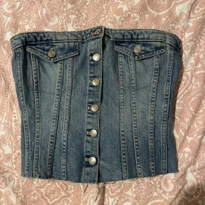 Jeans korsettopp med knappar - Snygg jeansfärgad korsettopp med knappar framtill och två fickor med lock och silverfärgade knappar. Perfekt för att skapa en cool och trendig look.