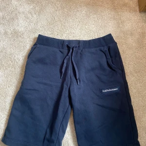 Mörkblå shorts från Peak Performance - Mörkblå shorts från Peak Performance med snörning i midjan och en bakficka. Mjukt material och logga på ena benet. Perfekta för avslappnade dagar eller träning.