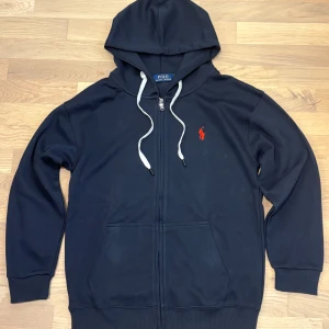Mörkblå hoodie från Ralph Lauren - Säljer en snygg mörkblå hoodie från Ralph Lauren med dragkedja och vit snörning. I princip perfekt skick garanterad att ge dig komplimanger! Storlek S och passar dig med S perfekt. 