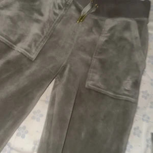 Gråa velourbyxor från Juicy Couture - Mysiga grön färgade velourbyxor från Juicy Couture med bred resår i midjan och dragsko. Byxorna har stora fickor med lock och guldiga knappar både fram och bak. Perfekta för en avslappnad och trendig look. Den här är guld detaljerad, och både ingår i setet, kofta o byxa, om inte du väljer köpa seprat, för 900 får du båda!
