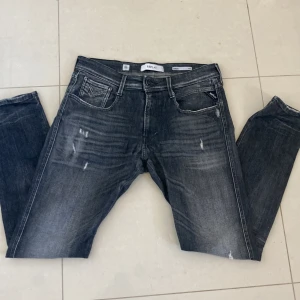 Replay Anbass slim fit jeans - Säljer ett par Replay Anbass jeans i slim fit med snyggt sliten look. Jeansen är mörkgrå med ljusa slitningar och klassiska fem fickor. Perfekta för dig som gillar en smalare passform och stilren design.